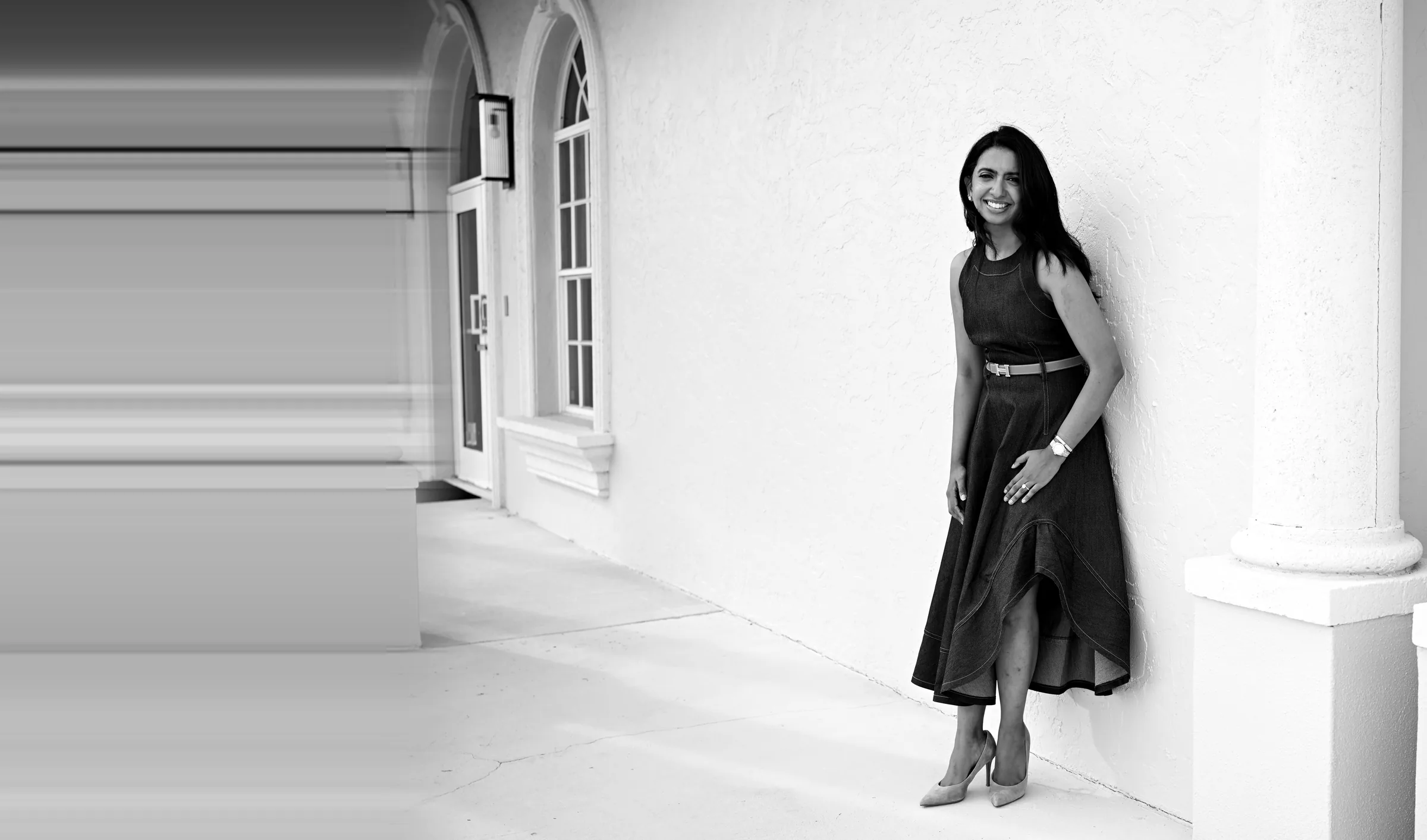 Dr. Meenachi Sellappan, cosmetic dentist, leaning against a white wall at Smile Spot Dentistry in Venice, FL — black and white portrait